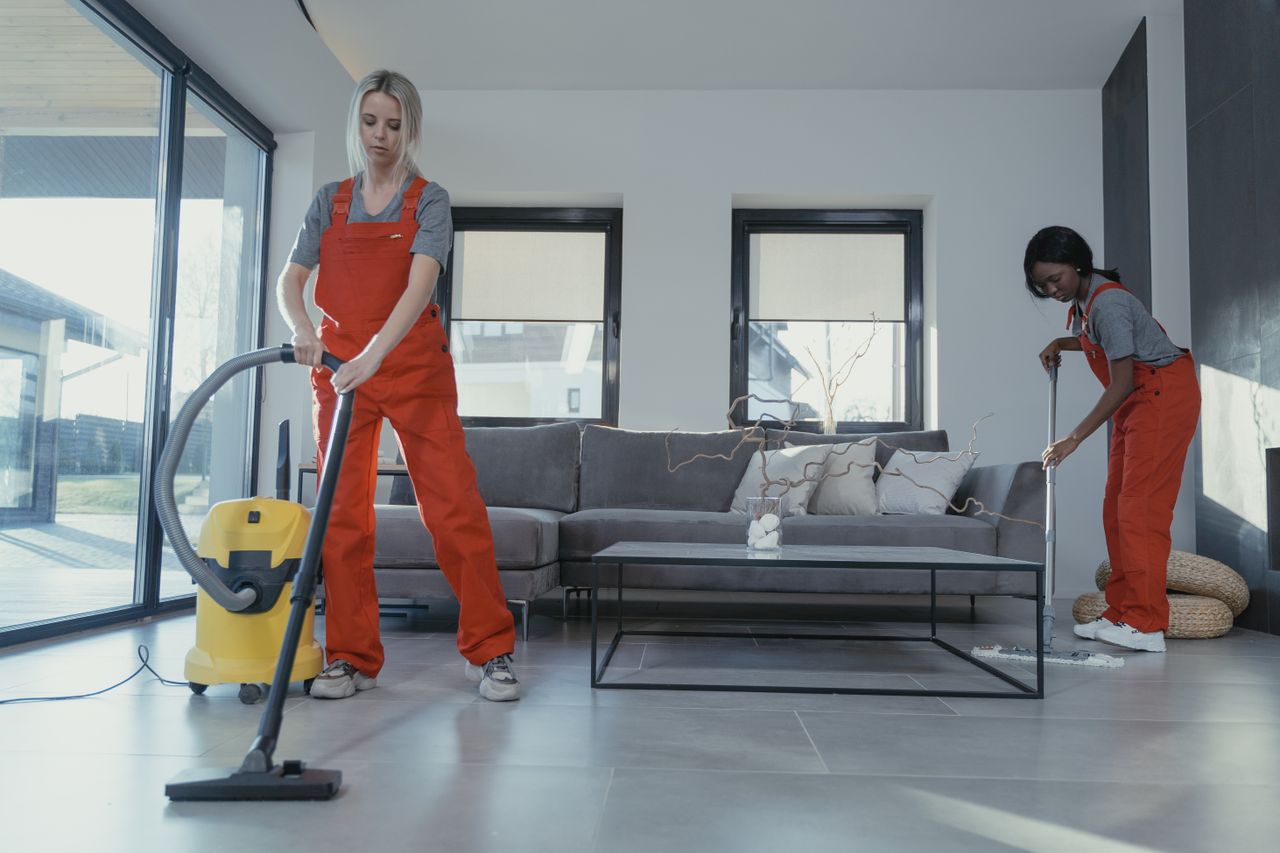 services-06 Two women in red overalls vacuuming and sweeping a modern living room.